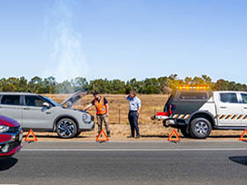 Breakdown service vehicle with flashing lights assisting a car on roadside, warning triangles placed.