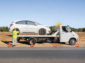 Tow truck with flashing lights loading a car on roadside, warning triangles placed for safety.
