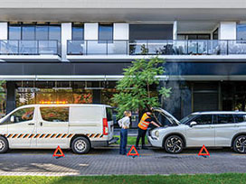 Breakdown service van with flashing lights helping a car on suburban street, warning triangles placed.