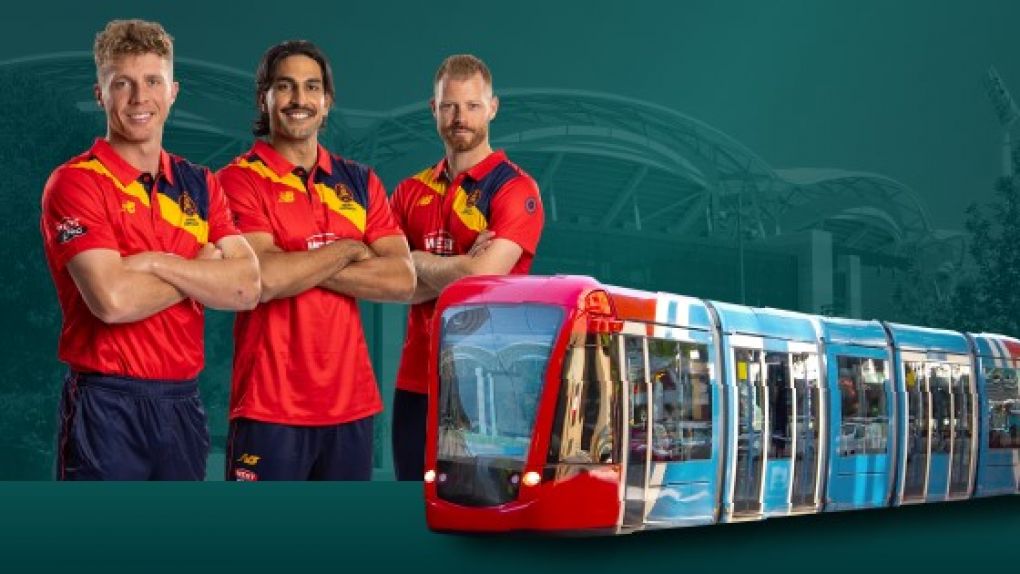 Three cricket players wearing the red, yellow and blue South Australian uniform stand next to an Adelaide Metro tram