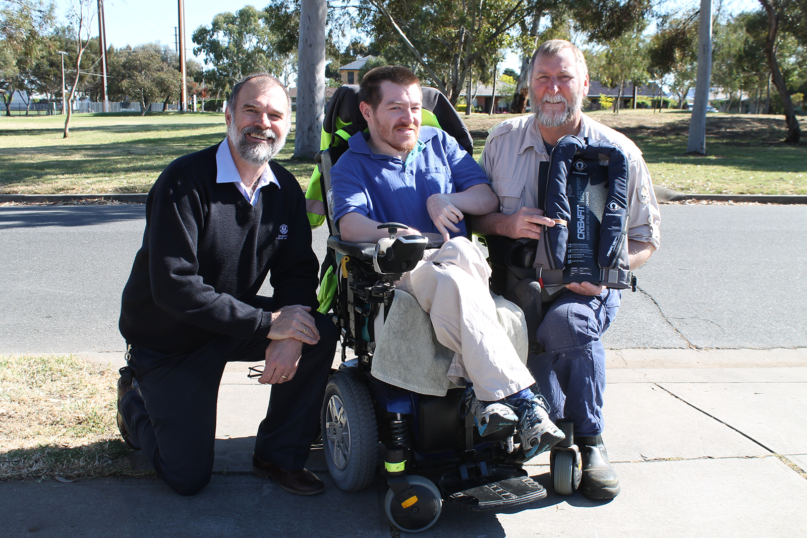 There are three men in the photo, all smiling. The man on the left is wearing a jumper with a government logo, the man in the centre is in an electric wheelchair and the man on the right is holding a lifejacket.