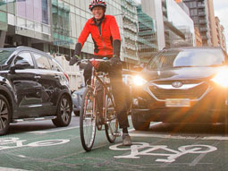 A cyclist wearing a red jersey is stopped in a green bike box. A car with headlights is stopped behind them.