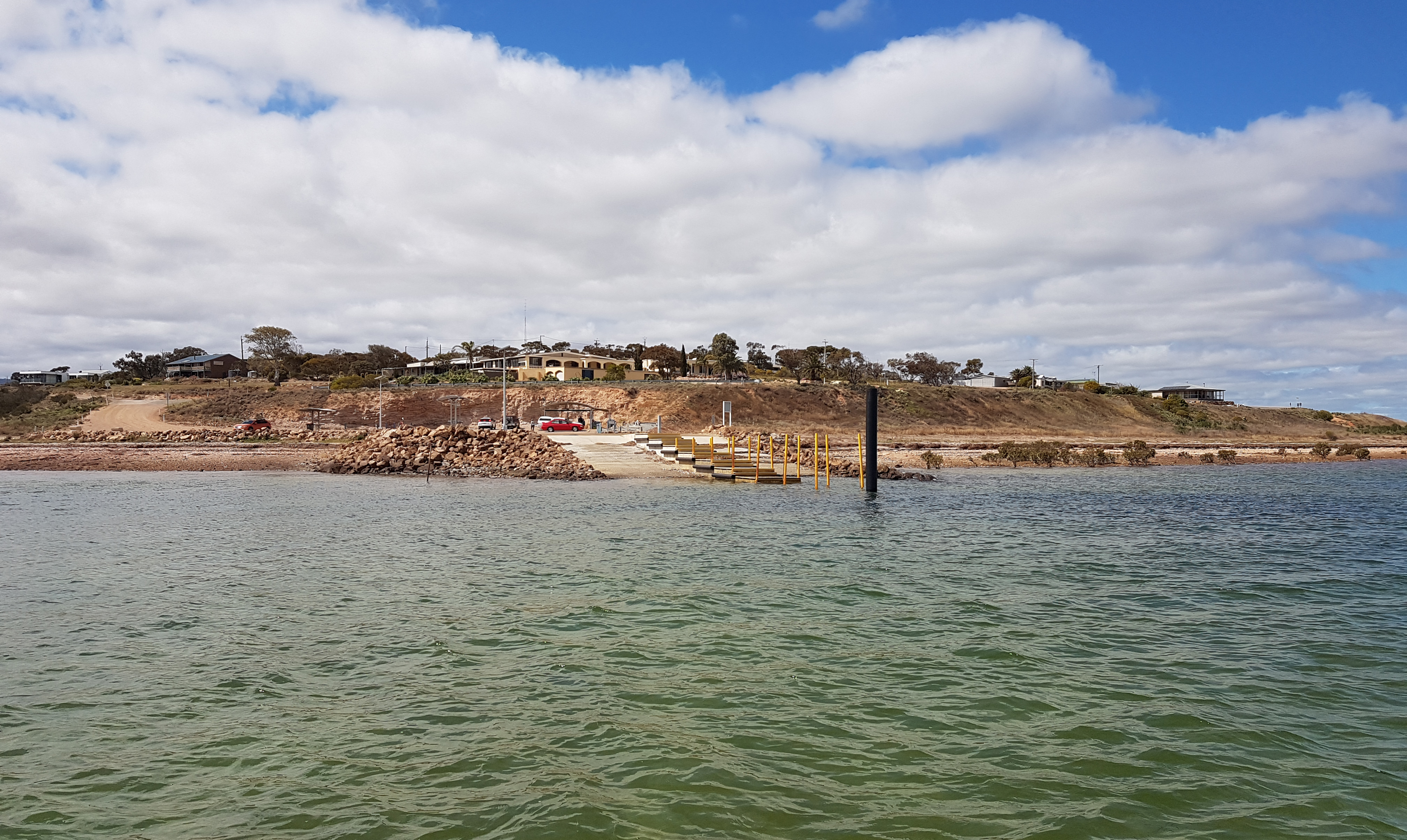 A picture of a boat ramp taken from the water. The ramp appears to be concrete with yellow poles on one side. There is a car park and homes on a hill in the background.
